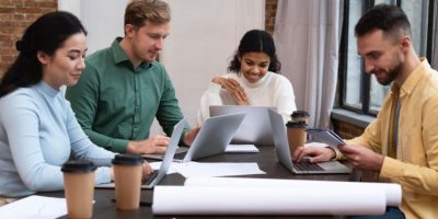 Four teammates working on laptops around a table, smiling as one checks his phone, representing digital marketing for small businesses collaboration.
