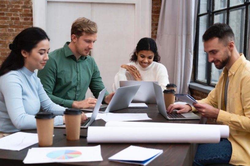 Four teammates working on laptops around a table, smiling as one checks his phone, representing digital marketing for small businesses collaboration.